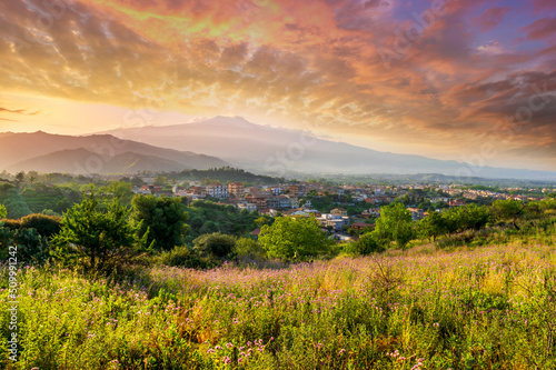 view from hill with golden grass and green bushes to a valley town with majectic mountains and scenic cloudy sunset on background © Yaroslav