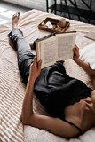  Beautiful young woman in  black silk pajamas reads a book lying on the bed in the bedroom.  