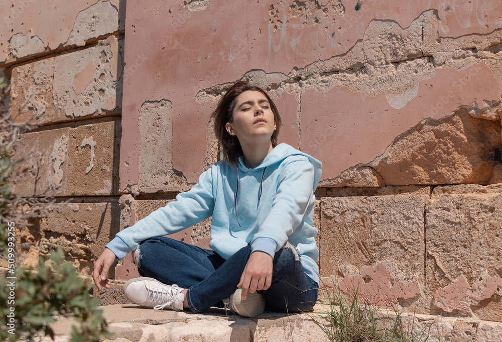 Teenager girl in light blue hoodie sitting on her haunches near a wall