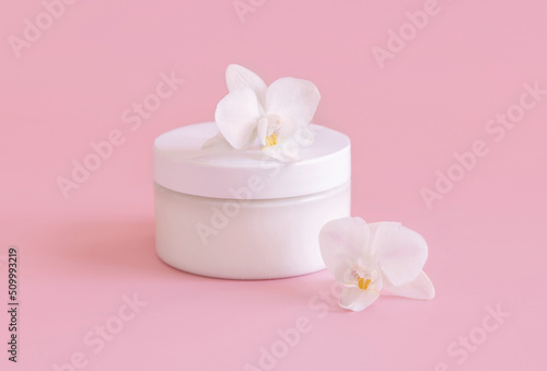 White cream jar near white orchid flowers on light pink close up. Mockup. Skincare product