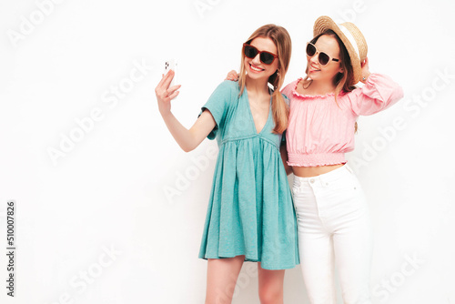 Two young beautiful smiling brunette hipster female in trendy summer dresses. Sexy carefree women posing near white wall. Positive models having fun. Cheerful and happy. In hats. Taking selfie