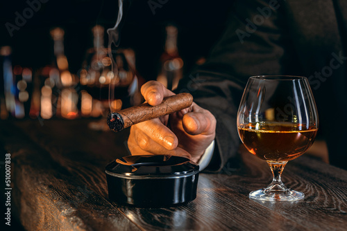 Man's hands with a cigar, elegant glass of brandy on the bar counter. Alcoholic drinks, cognac, whiskey, port, brandy, rum, scotch, bourbon. Vintage wooden table in a pub at night