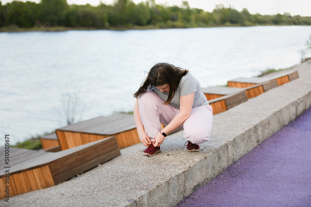 Overweight European teenage girl in tracksuit ties her shoelaces on her sneakers while jogging along on concrete embankment, Sports and teenagers, overweight teenagers