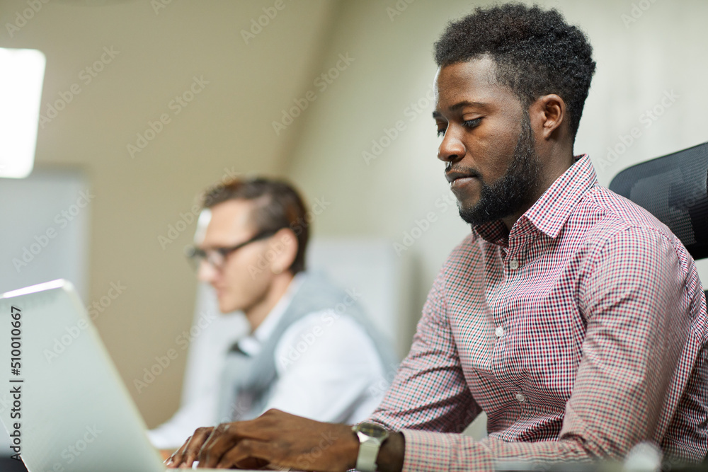 Concentrated young African-American programmer in checkered shirt sitting at table and typing on laptop Concentrated young African-American programmer in checkered shirt sitting at table and typing on laptop