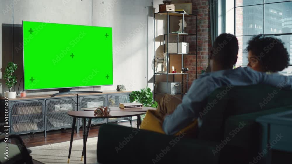 Black Couple Sitting and Watching TV with Green Screen Mock Up Display in Their Stylish Loft Apartment. Weekend Entertainment Concept. Shot From Back Black Couple Sitting and Watching TV with Green Screen Mock Up Display in Their Stylish Loft Apartment. Weekend Entertainment Concept. Shot From Back