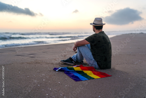 person sitting on the shore of the beach with rainbow flag watching the sunset photo