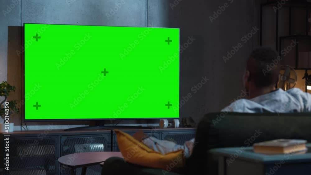 Black Man Watches Green Mock-up Screen TV while Sitting on a Couch at Home. Male Stream Evening Movie. Over the Shoulder View. Entertainment Concept Black Man Watches Green Mock-up Screen TV while Sitting on a Couch at Home. Male Stream Evening Movie. Over the Shoulder View. Entertainment Concept