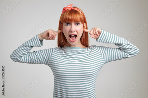 Indoor portrait of young ginger female posing over white wall plug her ears with fingers and screaming into camera