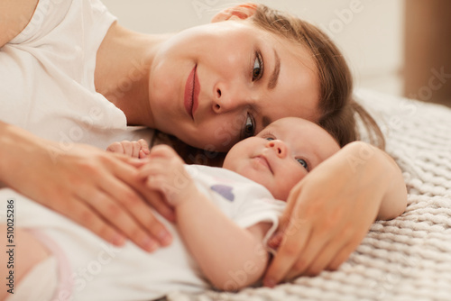 Happy pensive young woman lying on bed and looking at her adorable little child