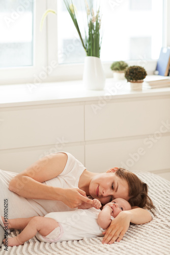 Calm young Caucasian woman lying on bed and looking at adorable little daughter squeezing her finger