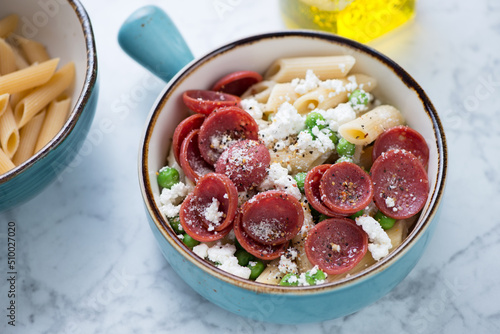 Penne pasta with crispy salami, ricotta cheese and green peas in a turquoise bowl, studio shot on a light-grey marble surface