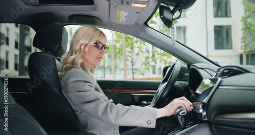 Female mature driver in formal clothes adjusting radio dial button control, gps or air conditioning while sitting behind the whel of car. Inside car view photo