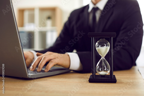 Close up of black hourglass with white trickling sand on table of businessman working on laptop. Hourglass at workplace of man in suit. Business, time and deadline concept.