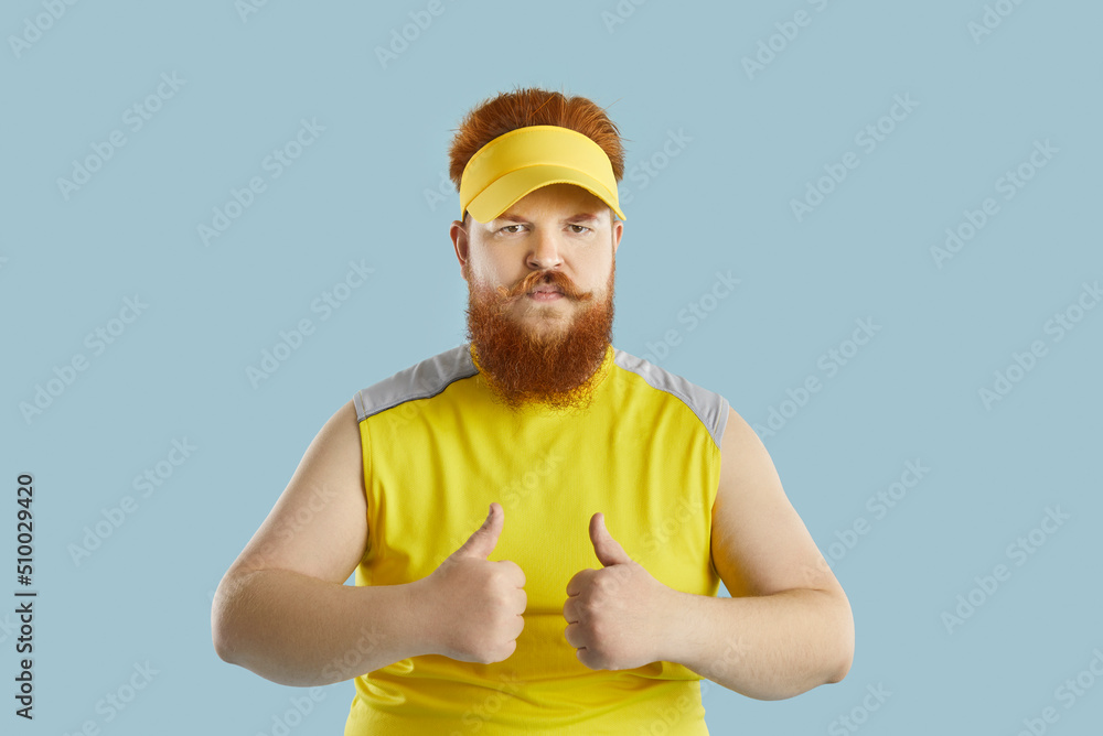 Portrait of serious redhead bearded man in sportswear isolated on blue studio background show thumbs up. Funny eccentric male client or customer give recommendation to offer or deal.