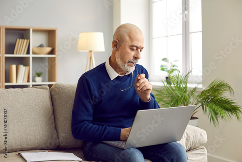 Man using laptop computer at home. Bald bearded senior man sitting on sofa in living room, working on modern laptop, writing email, thinking, looking at screen, considering ideas and biting glasses