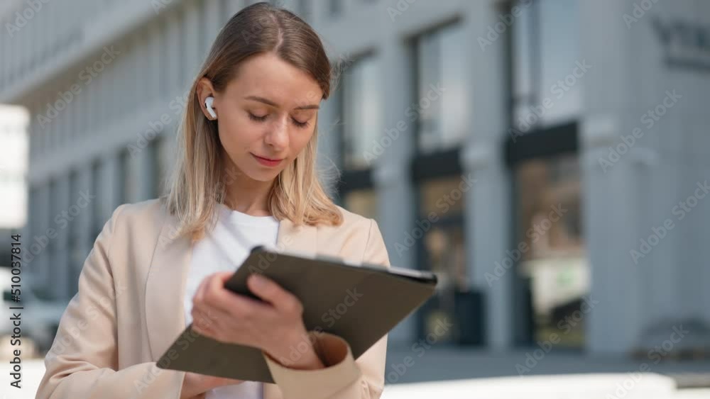 Professional businesswoman checking work mailbox on digital tablet outdoors. Focused caucasian woman in wireless earphones standing near office building. Professional businesswoman checking work mailbox on digital tablet outdoors. Focused caucasian woman in wireless earphones standing near office building.
