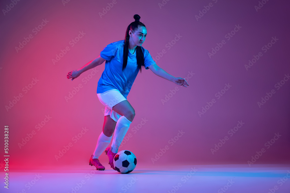 Dribbling. One sportive girl, female soccer player training with football ball isolated on purple studio background in neon light. Sport, action, motion, fitness