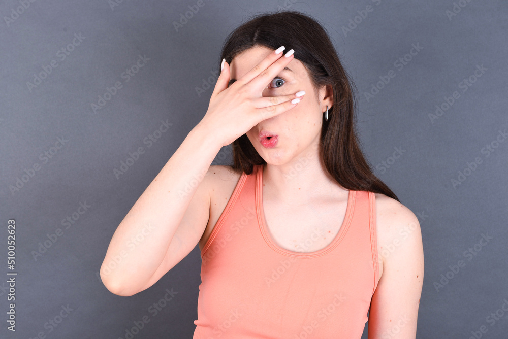 Caucasian woman wearing orange T-shirt over grey wall peeking in shock covering face and eyes with hand, looking through fingers with embarrassed expression. Caucasian woman wearing orange T-shirt over grey wall peeking in shock covering face and eyes with hand, looking through fingers with embarrassed expression.