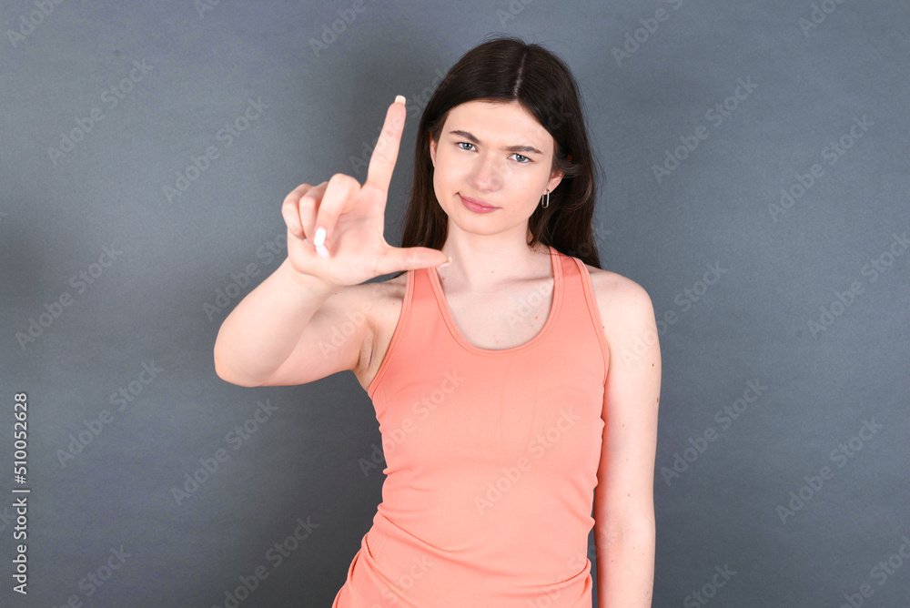Caucasian woman wearing orange T-shirt over grey wall making fun of people with fingers on forehead doing loser gesture mocking and insulting. Caucasian woman wearing orange T-shirt over grey wall making fun of people with fingers on forehead doing loser gesture mocking and insulting.