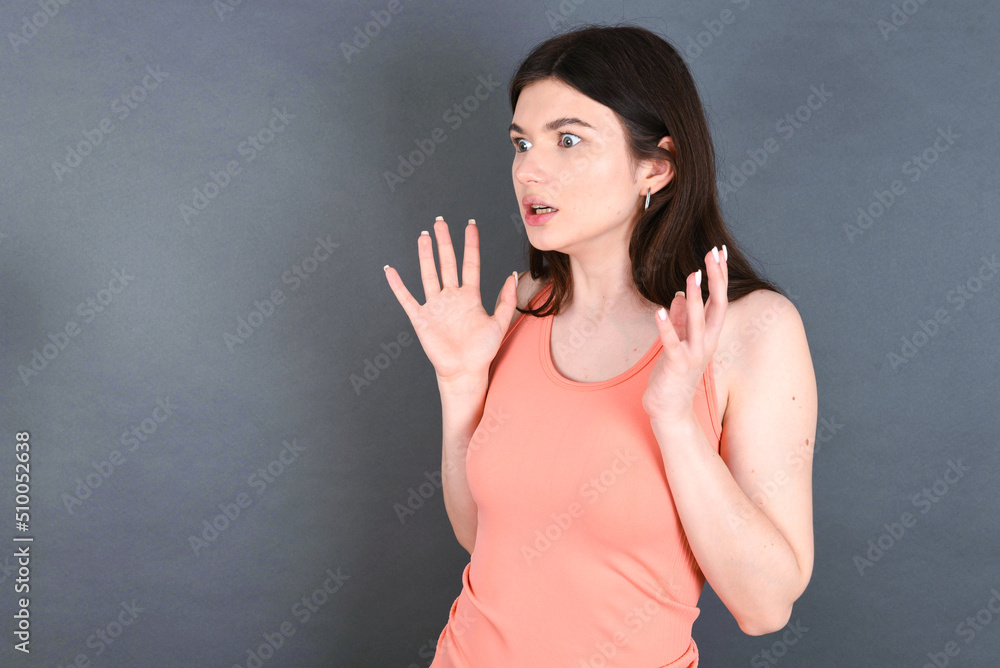 Caucasian woman wearing orange T-shirt over grey wall shouts loud, keeps eyes opened and hands tense. Caucasian woman wearing orange T-shirt over grey wall shouts loud, keeps eyes opened and hands tense.