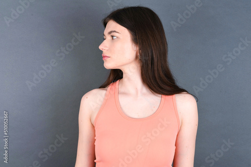 Close up side profile photo young beautiful Caucasian woman wearing orange T-shirt over grey wall not smiling attentive listen concentrated