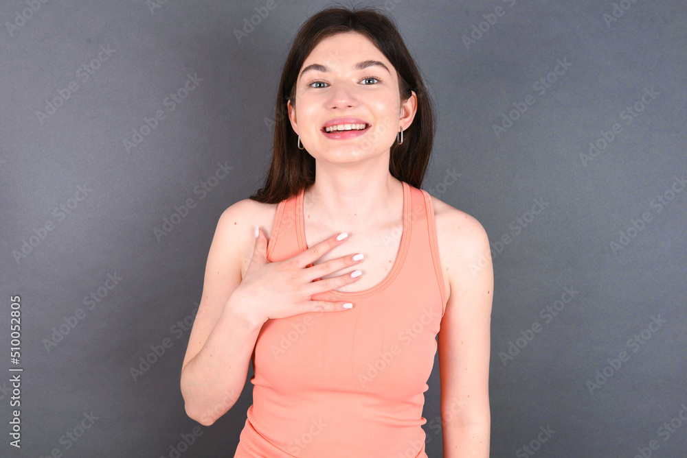 young beautiful Caucasian woman wearing orange T-shirt over grey wall smiles toothily cannot believe eyes expresses good emotions and surprisement young beautiful Caucasian woman wearing orange T-shirt over grey wall smiles toothily cannot believe eyes expresses good emotions and surprisement