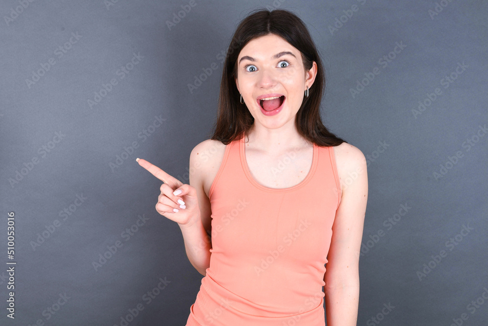 Caucasian woman wearing orange T-shirt over grey wall points aside on copy blank space. People promotion and advertising concept Caucasian woman wearing orange T-shirt over grey wall points aside on copy blank space. People promotion and advertising concept