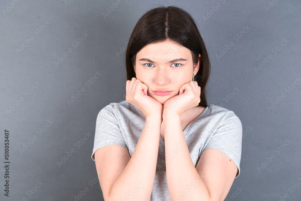 Portrait of sad young beautiful Caucasian woman wearing grey T-shirt over grey wall hands face Portrait of sad young beautiful Caucasian woman wearing grey T-shirt over grey wall hands face