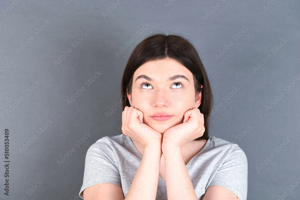 Portrait of sad young beautiful Caucasian woman wearing grey T-shirt over grey wall hands face look empty space Portrait of sad young beautiful Caucasian woman wearing grey T-shirt over grey wall hands face look empty space