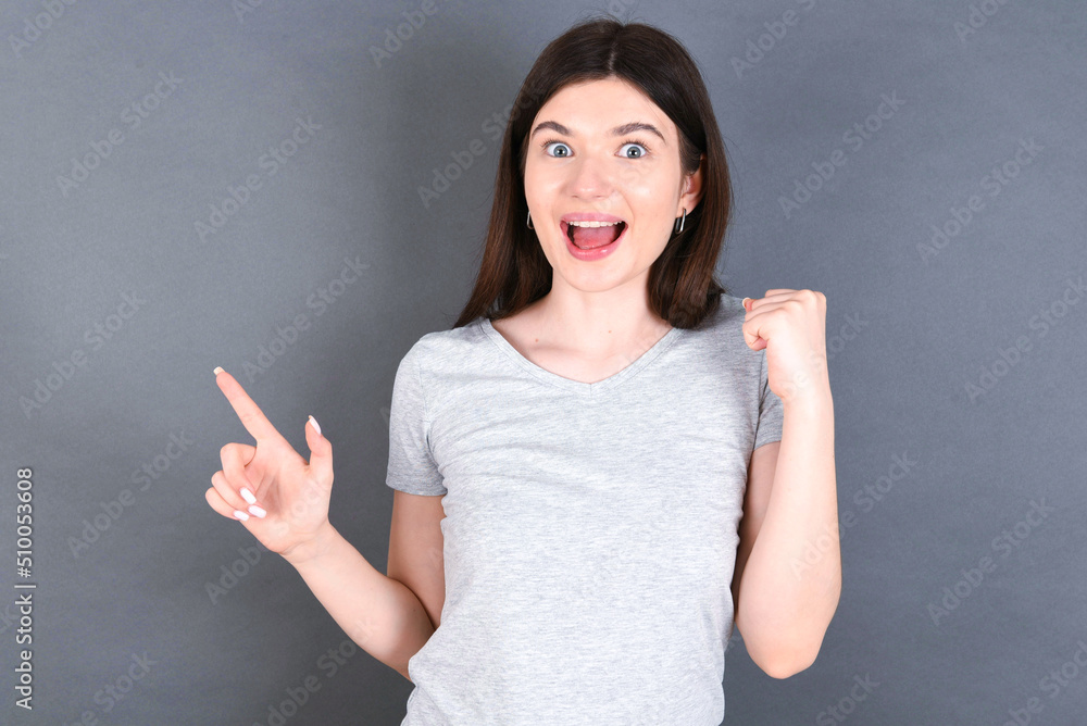 young beautiful Caucasian woman wearing white T-shirt over grey wall points at empty space holding fist up, winner gesture. young beautiful Caucasian woman wearing white T-shirt over grey wall points at empty space holding fist up, winner gesture.