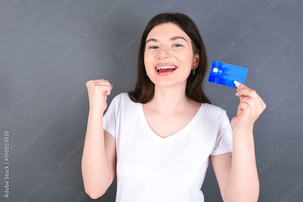 Young beautiful caucasian woman wearing white T-shirt over gray background holding a credit card, screaming and making winner gesture clenching and rising fist up. Young beautiful caucasian woman wearing white T-shirt over gray background holding a credit card, screaming and making winner gesture clenching and rising fist up.