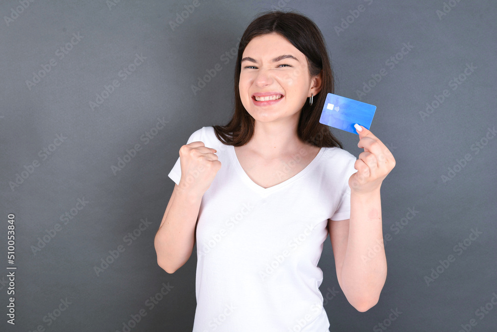 Young beautiful caucasian woman wearing white T-shirt over gray background holding a credit card, screaming and making winner gesture clenching and rising fist up. Young beautiful caucasian woman wearing white T-shirt over gray background holding a credit card, screaming and making winner gesture clenching and rising fist up.
