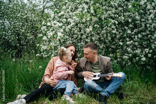 family dad mom baby daughter in the garden blooming apple trees, father playing the ukulele, scent of flowers outdoor nature © Svetlana Repnitskaya