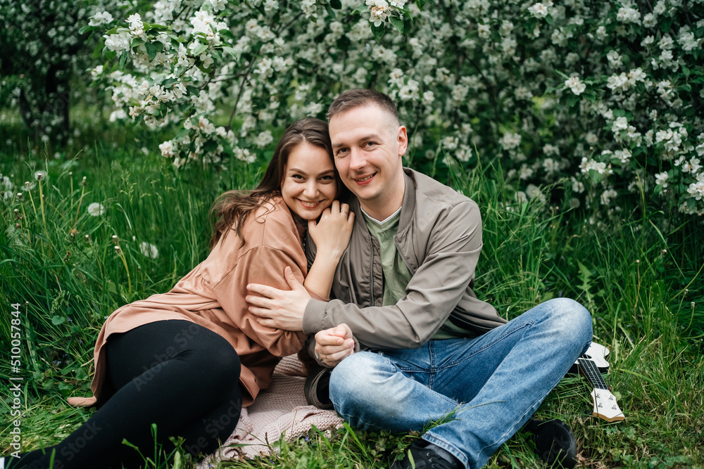 Fototapeta premium young couple in love a girl and a man sit on the grass of a flowering apple tree the aroma of flowers outdoors