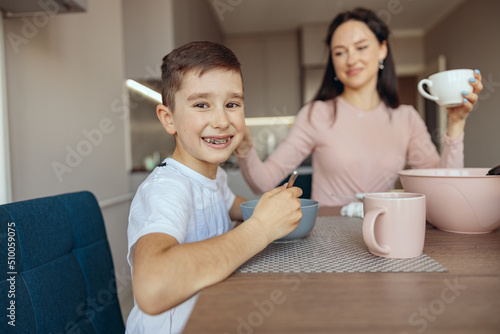 Portrait of joyful little Caucasian teen boy smiling and having breakfast with cereal. Kitchen.