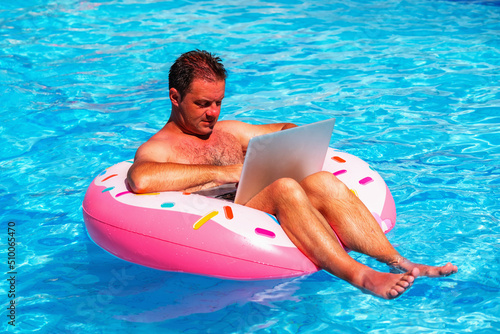 Portrait of businessman sitting on an inflatable circle in the swimming pool and working remotely with laptop. Horizontal image. © zwiebackesser