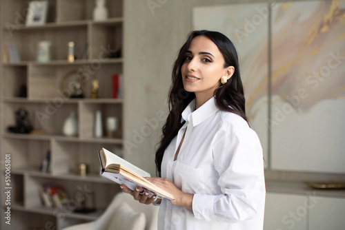 Woman in white shirt indoors in apartment interior holding book, education at home office, freelancer or student, personal development and self improvement, bookworm and passion in reading