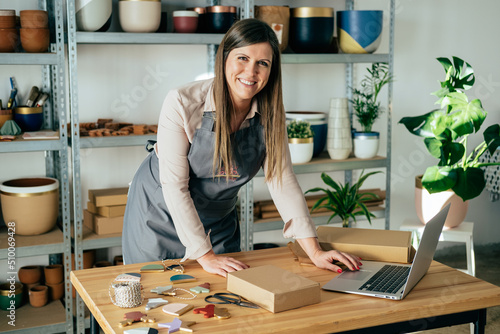 Portrait Of Smiling Woman In Apron Using A Laptop Computer And Preparing Christmas Ornaments Of Clay For Delivery At Her Store