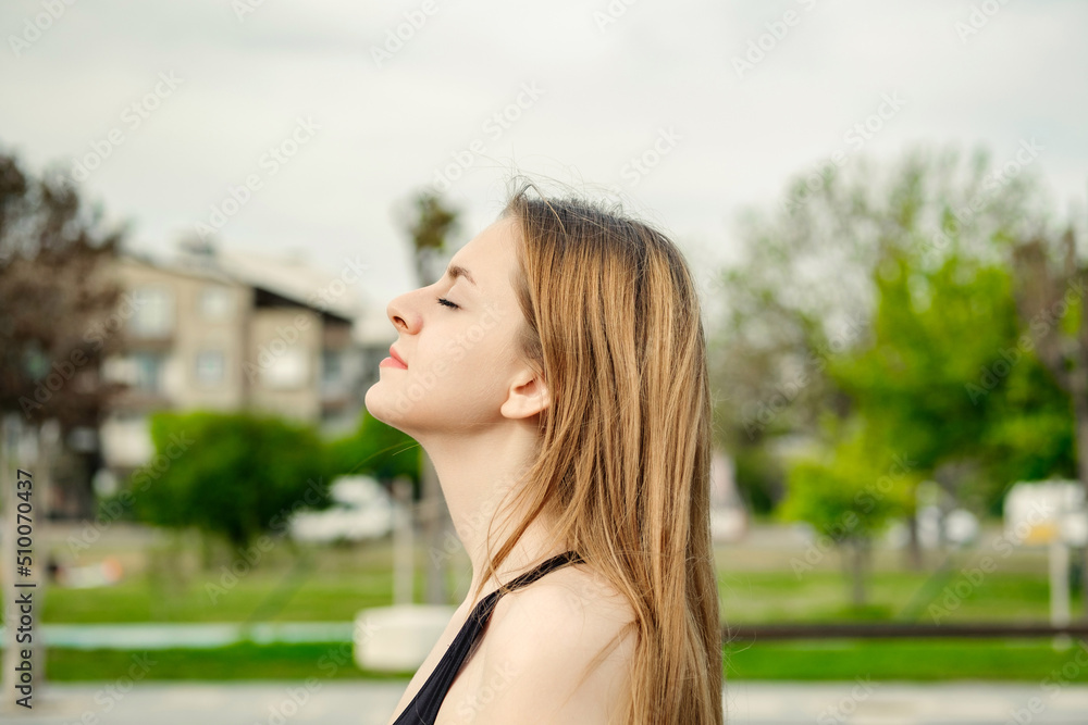 Brunette sportive woman wearing black sports bra standing on city park, outdoors relaxing with eyes closed, feeling alive, breathing, dreaming.