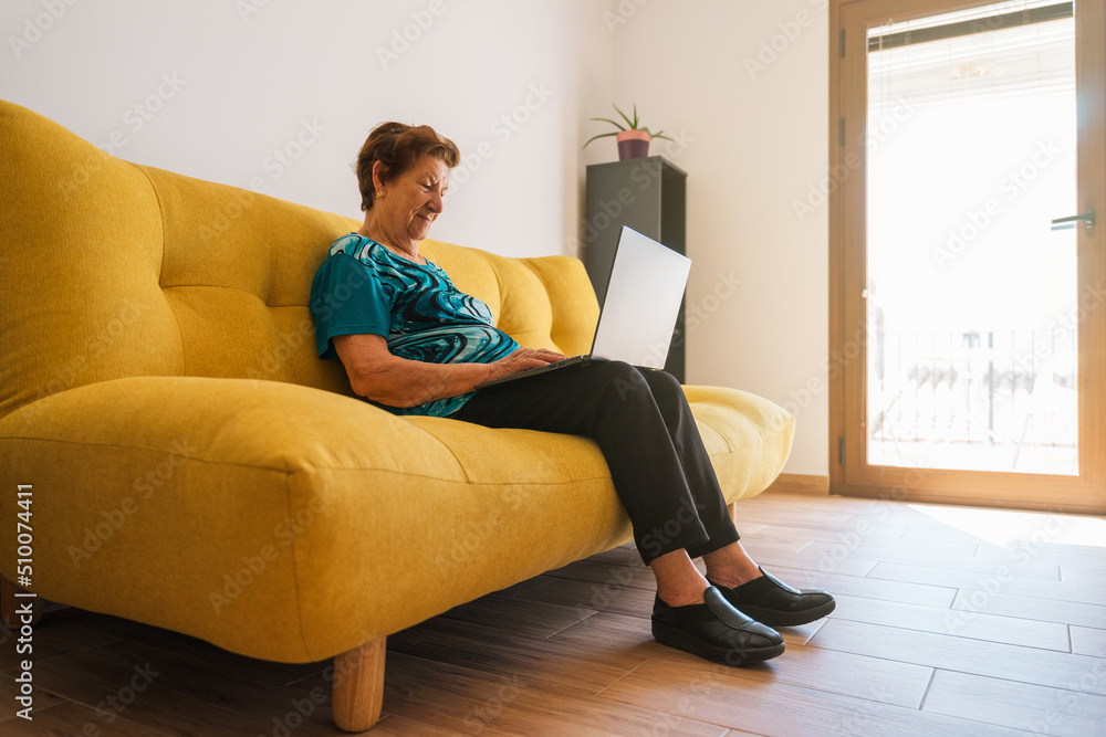 Open shot of an senior Caucasian woman using a laptop on a yellow sofa. She is wearing a blue T-shirt. Natural light in the room. Open shot of an senior Caucasian woman using a laptop on a yellow sofa. She is wearing a blue T-shirt. Natural light in the room.
