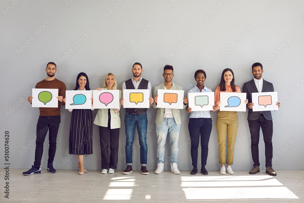 Obraz premium Everyone has their own opinion. Portrait of group of men and women holding white sheets of paper with empty colored speech bubbles on them. Multiracial people standing near wall and empty room.