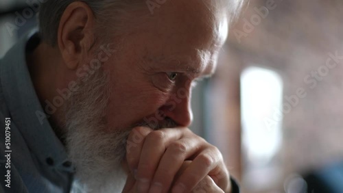 Close-up head shot of bearded gray-haired frustrated thoughtful senior adult man sitting alone at home thinking of lonely life. Sad depressed elderly male suffering from loneliness at nursing home.