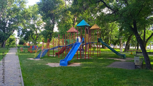 City Park. Playground. Colored elements. Without people. Green trees and lawn. Landscaping. Place for walking. Warm season.