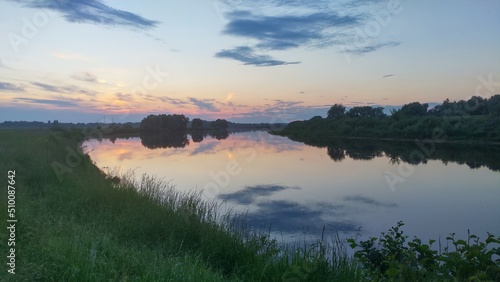 Late evening over the river, along the banks of which grass and trees grow. The sun has long gone down and the sky is painted in beautiful colors. Clouds are reflected in the calm water