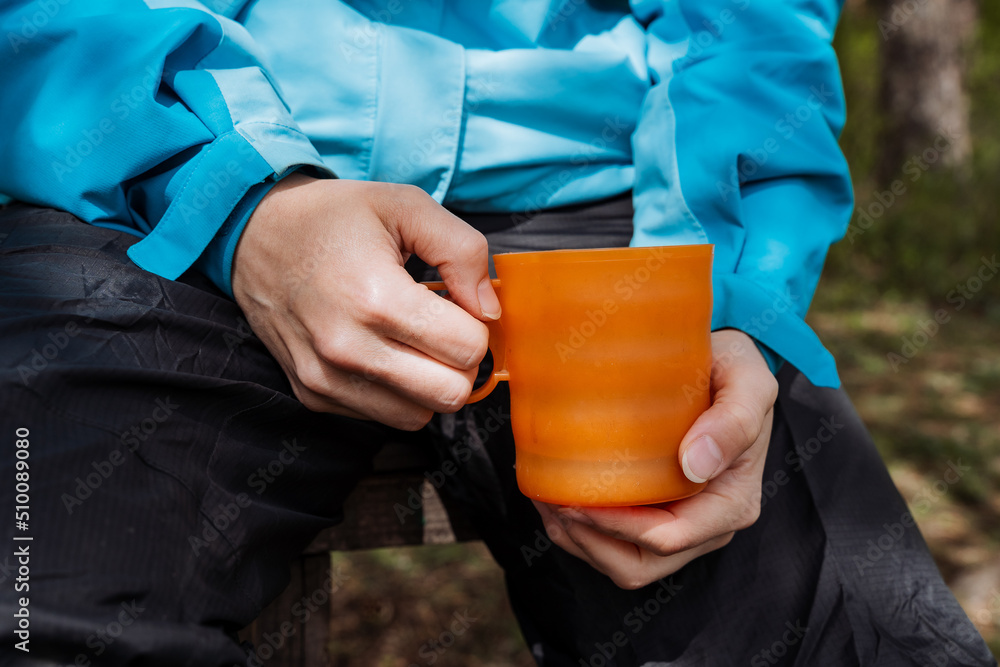 Women's hands hold a mug of coffee in nature, food on a hike to drink hot tea from a cup, an orange glass for drinks, a plastic mug eco product care for nature. Women's hands hold a mug of coffee in nature, food on a hike to drink hot tea from a cup, an orange glass for drinks, a plastic mug eco product care for nature.