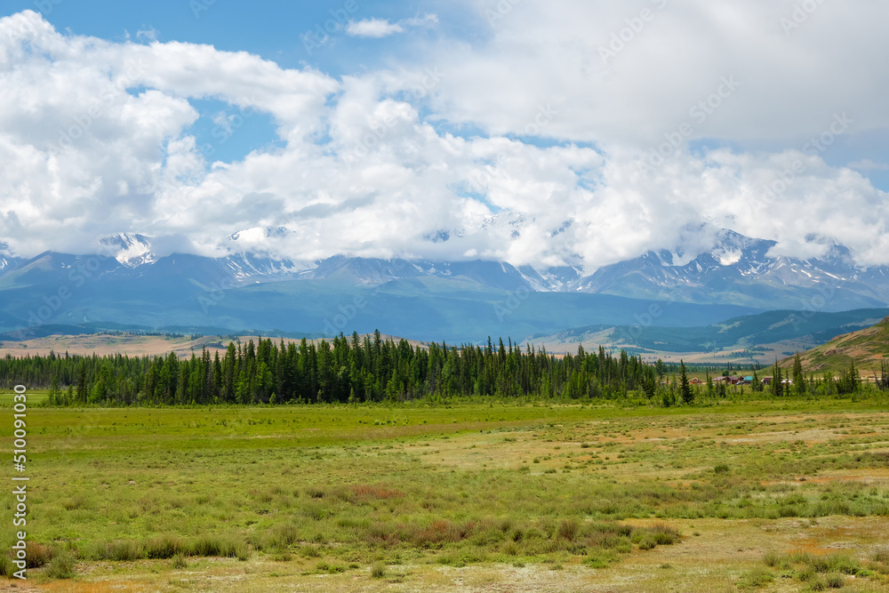 Remote mountain farm behind a cedar forest. Village life. A small remote village in Siberia. A green spring field in front of a forest on a mountain plateau against a cloudy sky. Agricultural land. Remote mountain farm behind a cedar forest. Village life. A small remote village in Siberia. A green spring field in front of a forest on a mountain plateau against a cloudy sky. Agricultural land.