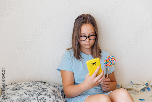 Teenage girl talks on the phone with a lollipop in her hand sitting on the bed photo