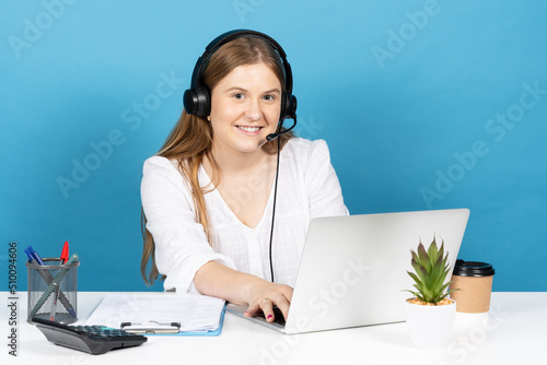 Young telephone or telemarketing operator working on laptop. Blonde woman looking at camera isolated on blue background.