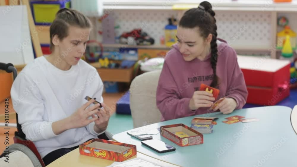 two girls with disabilities are playing a board game sitting at a large table. Rehabilitation of people with disabilities. two girls with disabilities are playing a board game sitting at a large table. Rehabilitation of people with disabilities.