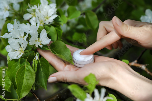 The moisturizing skincare cream in an open glass jar on woman's palm among the white flowers. Application of moisturizer